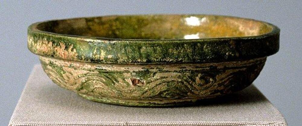Red earthenware, flat bottomed bowl with rounded sides and thick rim, molded with wave pattern to exterior wall, covered in amber and green lead glaze, with iridescence and calcification.
