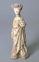 An earthenware figure of a tall thin woman dressed in sumptous robes that she has gathered in her left hand, her hair coiffed high upon her head.  It is covered in a white slip with traces of polychrome mineral pigment.