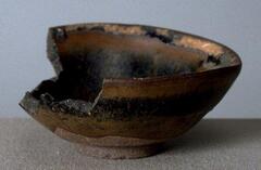 A conical bowl on a tall straight foot ring, covered in a dark brown-black glaze with russet hare's fur markings (兔毫盏 <em>tuhao zhan</em>).  There is significant loss to the side of the bowl.