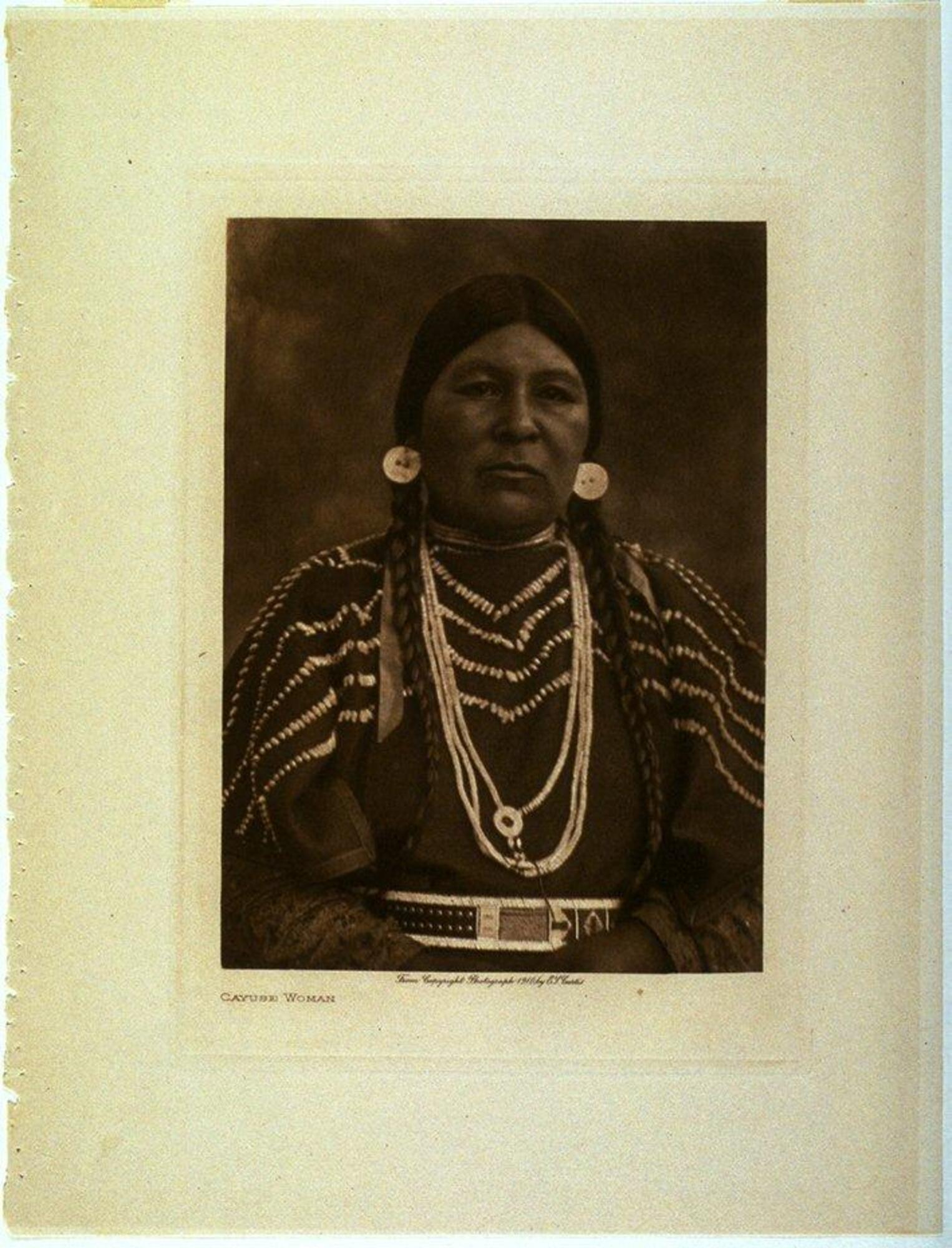 A portrait of a woman with long braids, large earrings, and beaded jewelry. She wears a shirt with geometric lines embroidered into the fabric, and an ornate belt.
