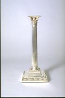 This silver candlestick, one of a pair, is in the shape of a fluted column topped by a composite capital composed of a double row of acanthus leaves and four volutes at the corners. The column rests upon a stepped base made of four squares of diminishing size, the first and fourth of which are marked with a pattern of strigillations.