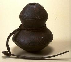 Vessel with two bulbous sections and a tapered middle section with a small opening at the top. There is a braided fiber string wrapped around the tapered mid section with a piece of wood attached to the end of the fiber. The bulbous portions of the vessel are decorated with geometric patterns.