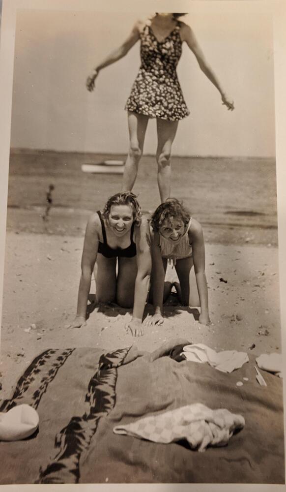 Three women form a pyramid on a beach. The two women on the bottom are on their hands and knees, while the third woman stands on their backs with her head out of frame.&nbsp;