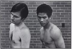 Two prisoners, shirtless in a line. There is a brick wall behind them.