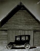 This image depicts a driverless Model T Ford automobile sitting in front of a wooden barn.