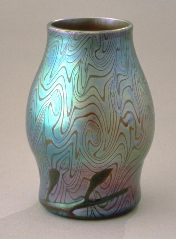 This iridescent blue and green glass vase has surface decorations that resemble peacock feather &quot;eyes&quot;. Along the bottom is a raised design of a branch or leaves that was added to the vessel late in its creation.