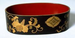 An oval tray with deep sides. Inside is a red lacquer with gold floral designs in lacquer on the outside against a dark brown background. A small geometric crest appears on the outside. Part of a bridal trousseau.