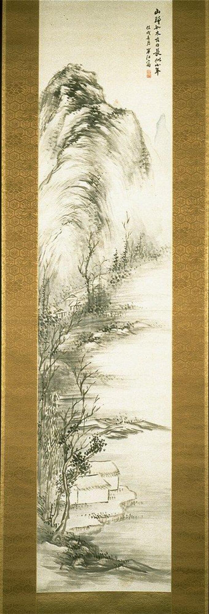 There are mountains in the background that run along a river. These mountains slowly turn into trees, many without leaves. There are two houses along the waterside, partially hidden behind the trees. There is an inscription and two seals in the upper right corner. The hanging scroll is bordered in gold.
