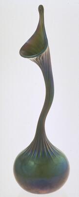 This elongated vase has a small, round base and an undulating, swan-like attenuated neck that ends in a flaring mouth. The length of the neck of the vessel has ridges and the surface has a dark iridescent quality of green and blue