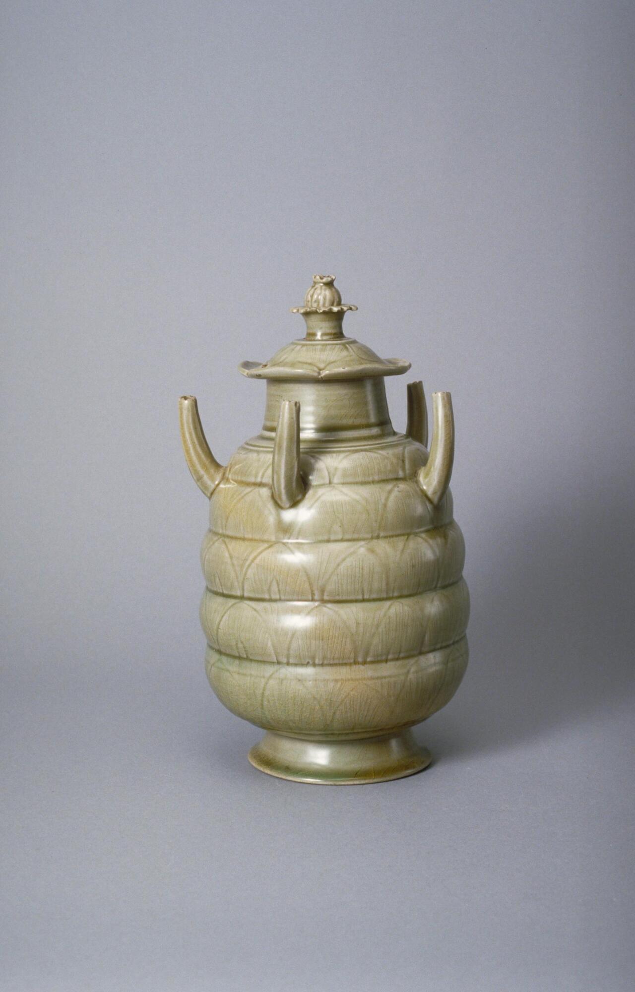 A buff stoneware jar rising up from a tall foot ring in an elongated globular body with lobes tapering towards the mouth. This jar has incised decoration, and five tubes evenly spaced and protruding upwards from the shoulder. The mouth is covered with a high domed lid, incised, and topped with a lotus bud finial. The jar is covered in an olive green celadon glaze.