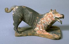 A gray earthenware sculpture of a crouching dog on a platform, resting on his two front paws, mouth open and pointed ears, long tail with curled end, with polychrome mineral pigments. 
