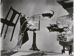 A studio image of artist Salvador Dali in a surrealistic scene. Dali jumps in the air as furniture hovers around him, and three cats fly into a stream of water that splashes across the scene.