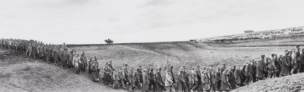 Wide-angle photograph of soldiers marching through a hilly landscape. Men on horseback and a spattering of buildings dot the horizon line.