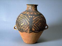 A light reddish-buff earthenware <em>guan (</em>罐) jar, with a wide globular upper body and conical lower body on a flat base, and a tall narrow neck with an everted rim. There are two diametrically opposed lug handles at the waist. The upper half of the body is painted with black pigment to depict four, six squared spirals made up of lines containing small circles, divided by thick vertical lines, confined between solid band borders with a lobed line border below. Around the shoulder, contained by two solid black lines, is a band of black squares divided by a central black vertical line, alternating in their position to touch the top or bottom solid black lines containing them.   Surrounding the neck are four bold vertical lines made up of wide and densly spaced zigzags. 
