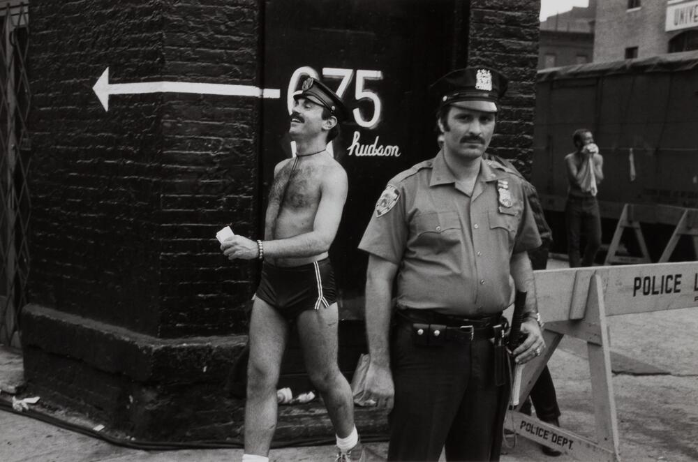 An actual police officer looking at the camera and an actor playing a police officer walking behind him.