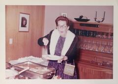 A woman in a fuzzy jacket and cat-eye glasses serving food from a buffet tray onto a plate in her hand. She looks at the camera with a surprised expression. There is a cabinet full of glassware in the background.