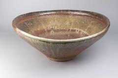 Large bowl with wide and asymmetrical rim and natural ash glaze. The bowl appears to be orange on the outside, and multi-colored on the inside.