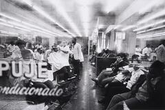 View inside a crowded barbershop; the reflections of lights both inside and dart back and forth from the numerous windows and mirrors throughout the image. 