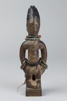 Standing male figure on a square base with hands at the sides. There is a geometric pattern carved in the abdomen and two strings of black beads around the waist. There are also two strands of beads and a brass bracelet on each wrist and two strings of multi-colored beads around the neck. There is a triangular pendant around the neck and much of the face has been worn smooth. The hair is in a conical shape coated with blue pigment. 