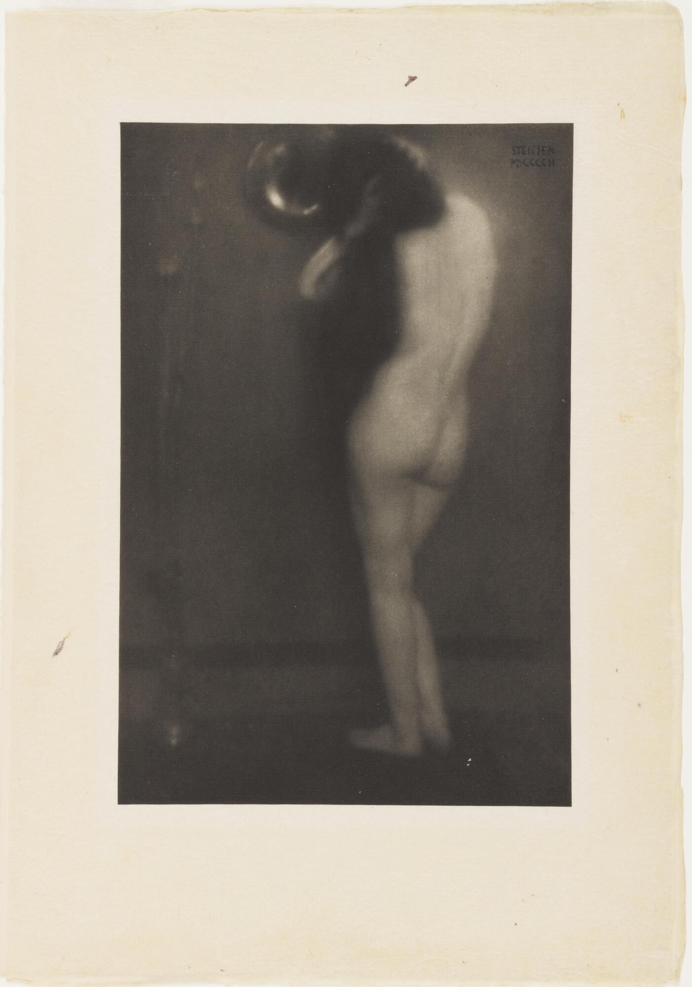 A nude woman stands with her back to the viewer. Her hands rest on her long dark hair as she gazes into a small, round mirror. 
