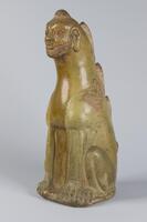 This stoneware figure of an anthropomorphic form consists of a lion-like body and paws, wings, human face with beard, and central flame-like horns and spikes extending down the back. The figure is covered in a straw-colored or light greenish-brown glaze. 