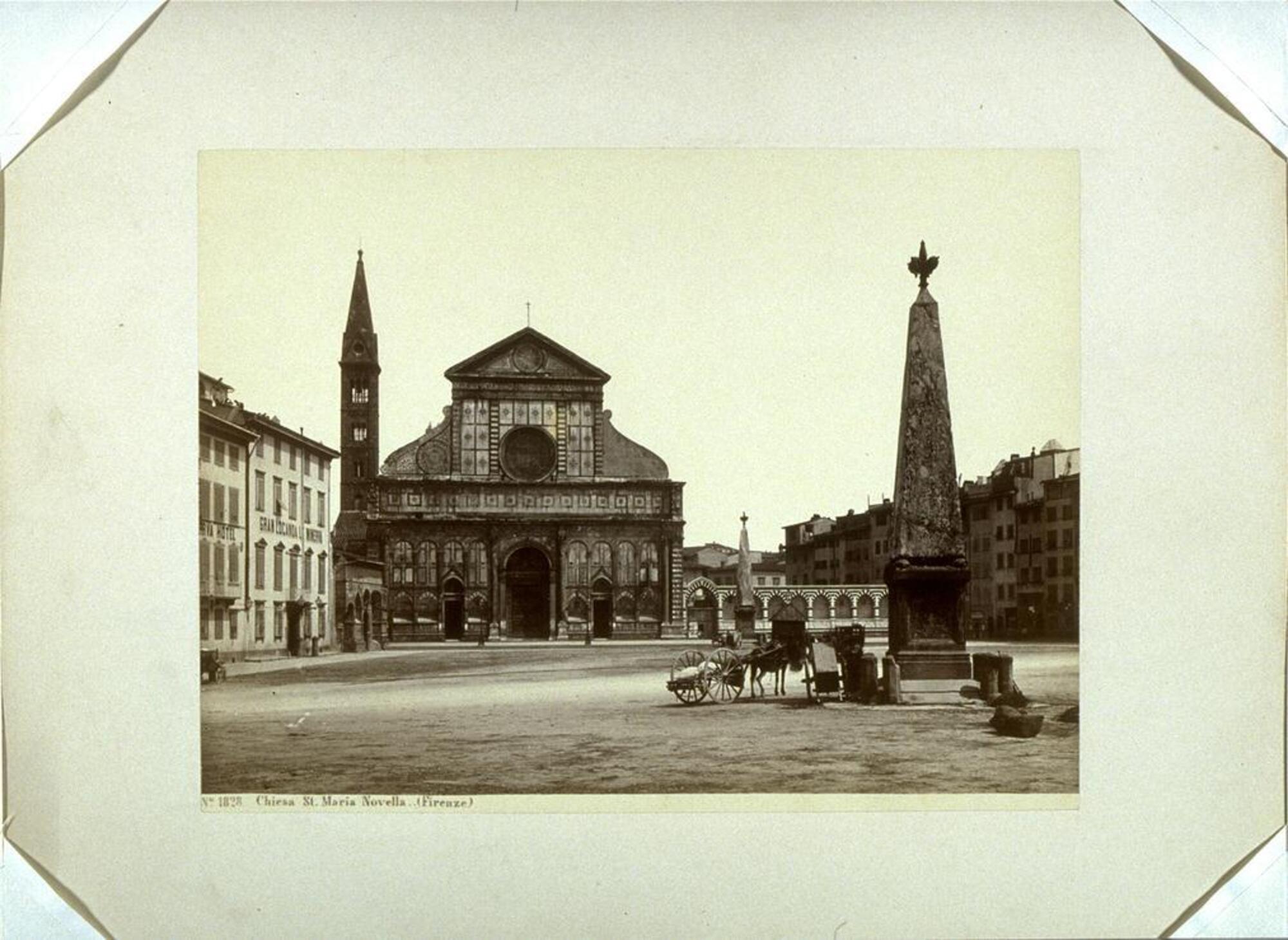 This photograph depicts a view of a church, before the church is a wide open piazza with two obelisks standing in its center. Next to one of the obelisks is a donkey cart.