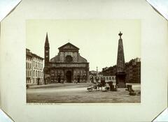 This photograph depicts a view of a church, before the church is a wide open piazza with two obelisks standing in its center. Next to one of the obelisks is a donkey cart.