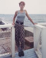 Woman standing on a deck wth the sea in the background. She faces the camera and wears one earring in the shape of a cross.