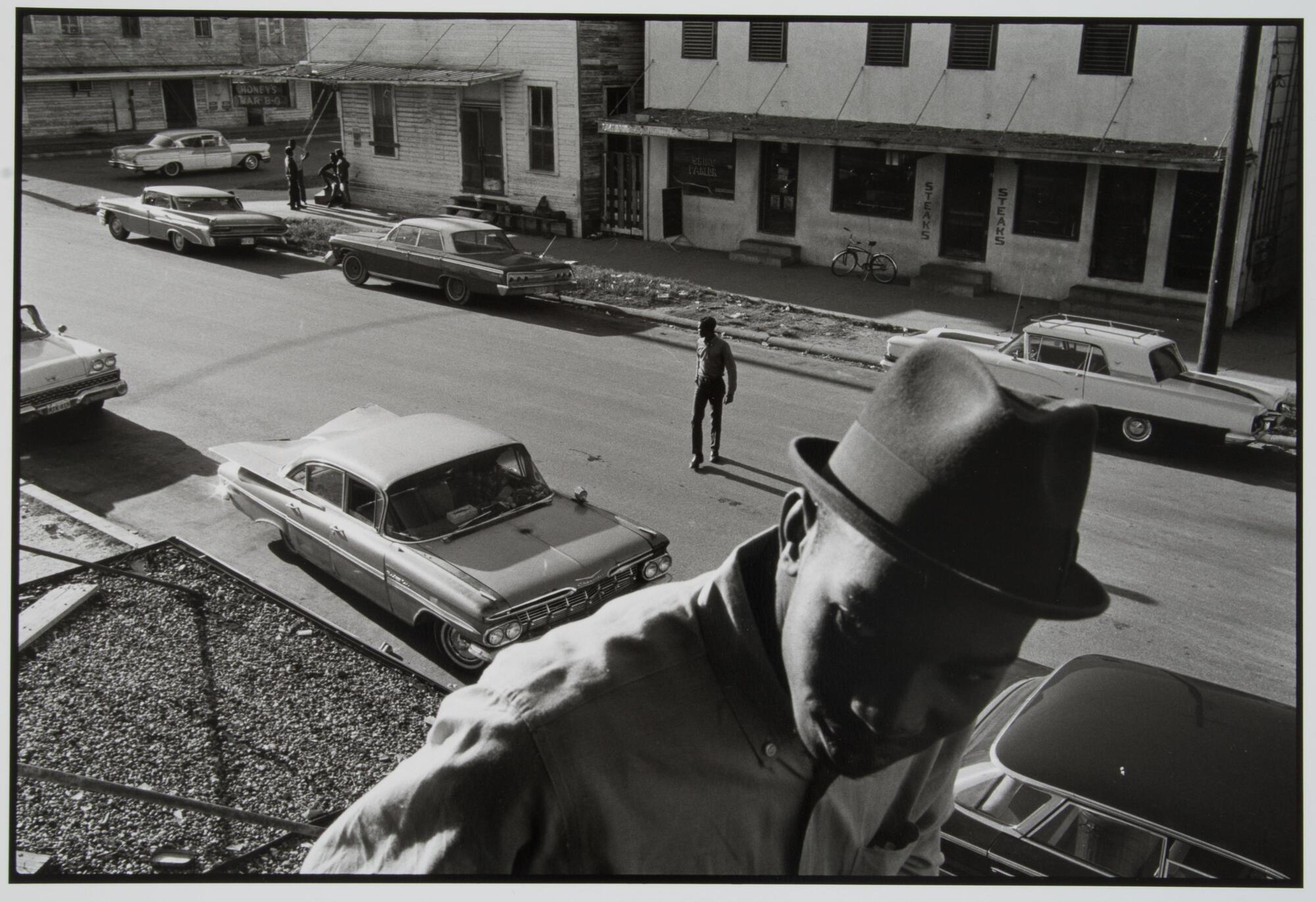 A photograph taken from a roof. In the foreground, an African American man stands on the roof wearing a jacket and hat. A street lined with cars and a lone pedestrian are visible below.