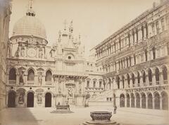This photograph depicts a view of a courtyard inside the walls of an ornate, palatial building. The figures of two men stand around a well-head, while a third man in uniform watches from a distance.