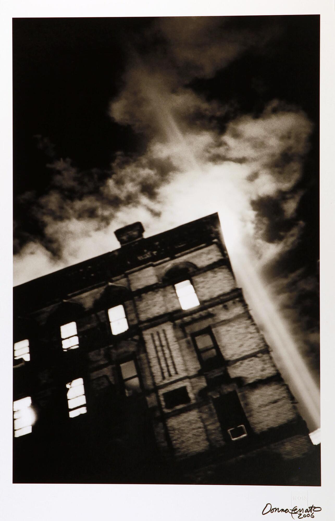 A black and white photo of a multi-story building.  Two spotlights shine straight up into the clouds just to the right of the building.