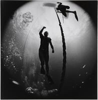 A diver wearing swimming fins is seen from below. A rope to his right rises to the surface, reaching another diver above. A black circle frames the entire scene, extending to the edges of the photograph.