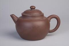 Stoneware teapot with round handle, smooth body, lid, and short spout. The lid contains two ridges and a small rounded knob at top center. Below the lid on the body is also a concentric ridge.