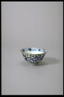 A petite cup with an abstract floral design in blue, white, and gold covering the outside of the cup and inside rim. The outside rim is gold and slightly everted. It has a large foot with blue stripes.