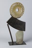 This sculpture has a rectangular metal base supporting a gray stone and bronze rod, which together support a curved rectangular bronze element. A yellow glass wheel with a square radial and protruding dimples tops the work. 