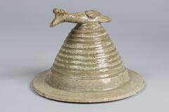 This ribbed, cone-shaped,&nbsp;porcelain lid has a wide flange topped with a bird-shaped finial. It is covered in a white glaze with a bluish green tinge.