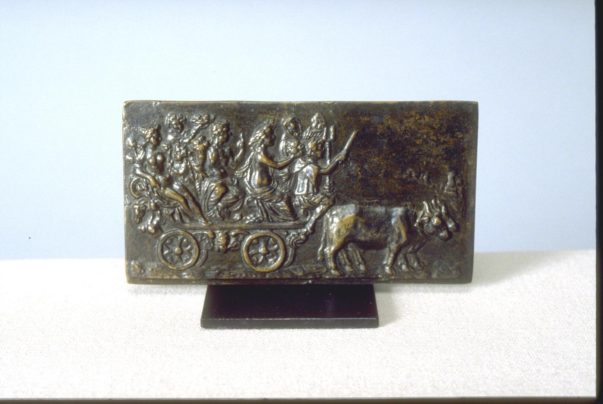 This bronze plaque features a pair of oxen pulling a four-wheeled wagon with six passengers. The seated driver holds a rod in his right hand and a two-tined fork in his left. An older figure holding a flaming vessel stands behind him, followed by a seated figure wearing classical drapery and a laurel wreath. The next figure, seated in the middle of the cart, is an older female, nude to the waist, who holds a cornucopia full of fruit and raises her left hand to point skyward. Two smaller standing figures appear next, one holding a bowl of fruit. The final figure is a reclining female nude holding a flower in the crook of her right arm.
