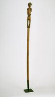 This smooth, wooden Pende staff features a finial depicting a standing, male figure bearing half-moon eyes, a terraced coiffure with decorative motifs, an elongated trunk, angular curves at the elbows and buttocks, and hands placed in front of the stomach below the navel. The feet are carved as one piece (forming the base of the finial), with small incisions for the individual toes. The left arm has been damaged.  