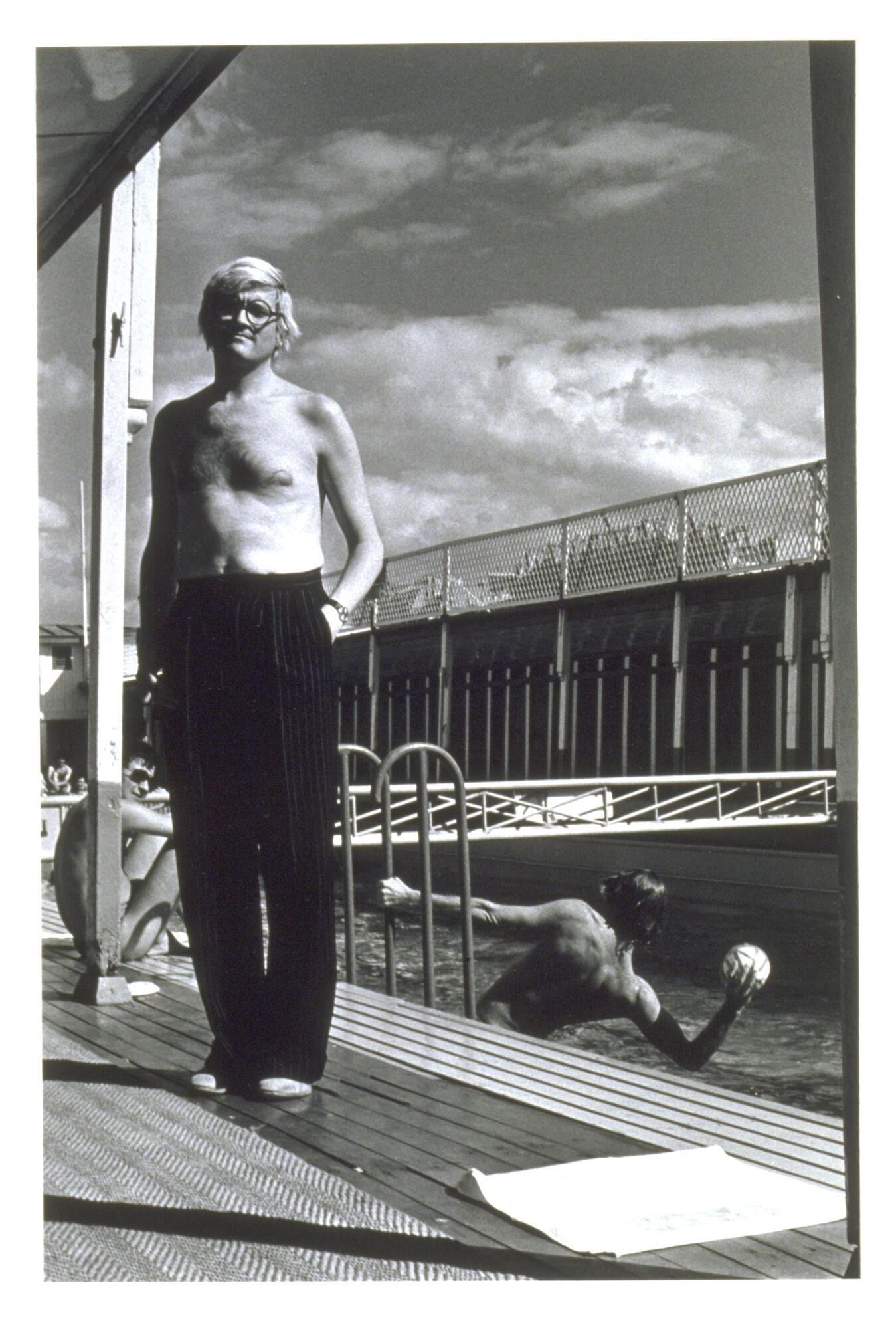 A photograph of man standing shirtless in front of a pool where others sunbathe and play water sports. 