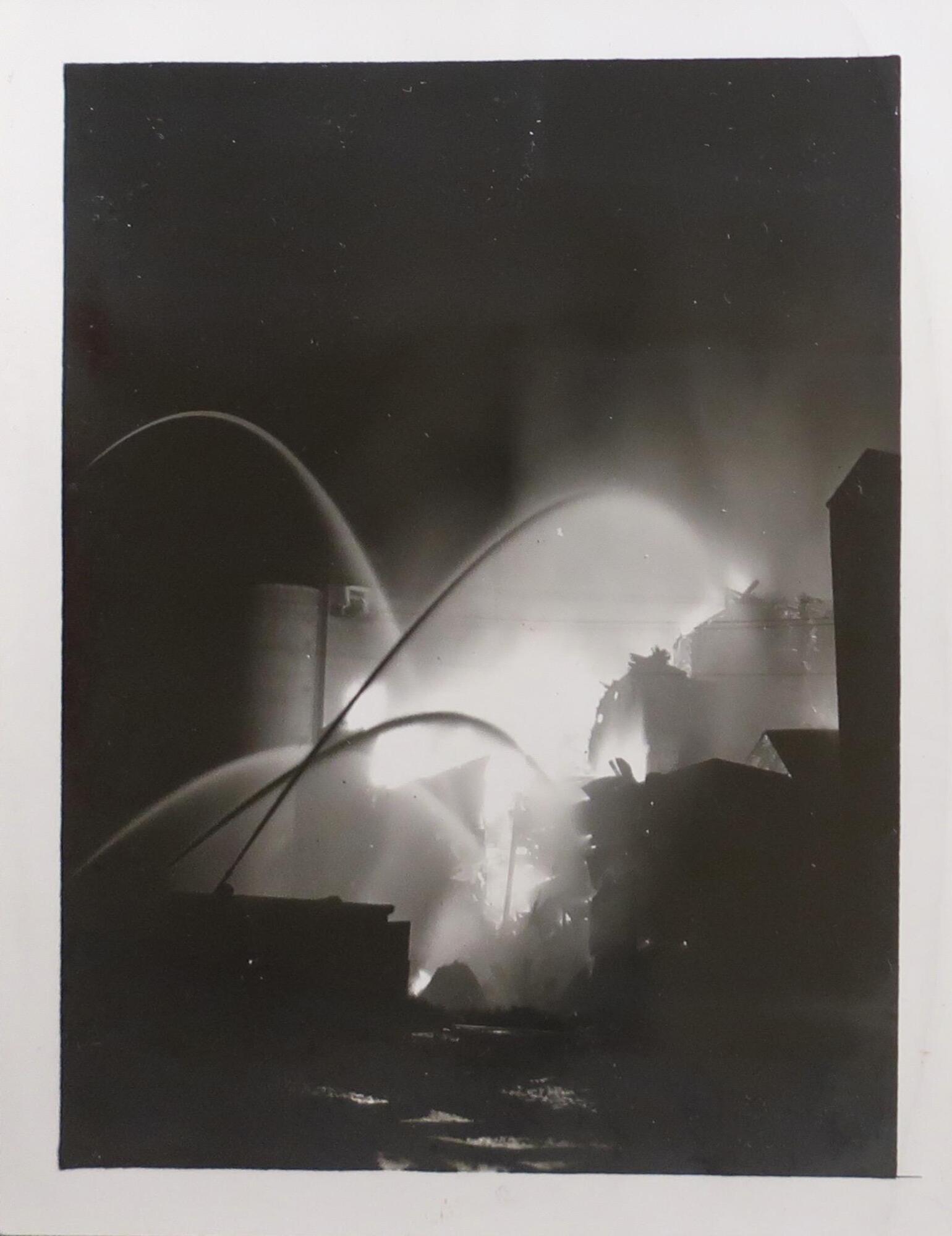 A burning building viewed from a distance. Four large jets of water arc towards the center from the left side of the image. The edges of the image are dark with silhouettes of buildings on the right side.