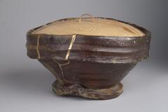 The bottom of this vessel is a brown glaze and the top is made of curved wood that snugly fits inside. The lid has a small handle on top.