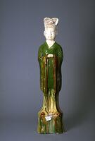 A standing civil official of earthenware, wearing a long robe with wide sleeves and with hands clasped in front of his chest.  He is wearing a tall hat divided into two lobes and standing on a tall pedestal.  With the exception of the head and hands, the figure is covered in runny amber, green and cream glazes. 