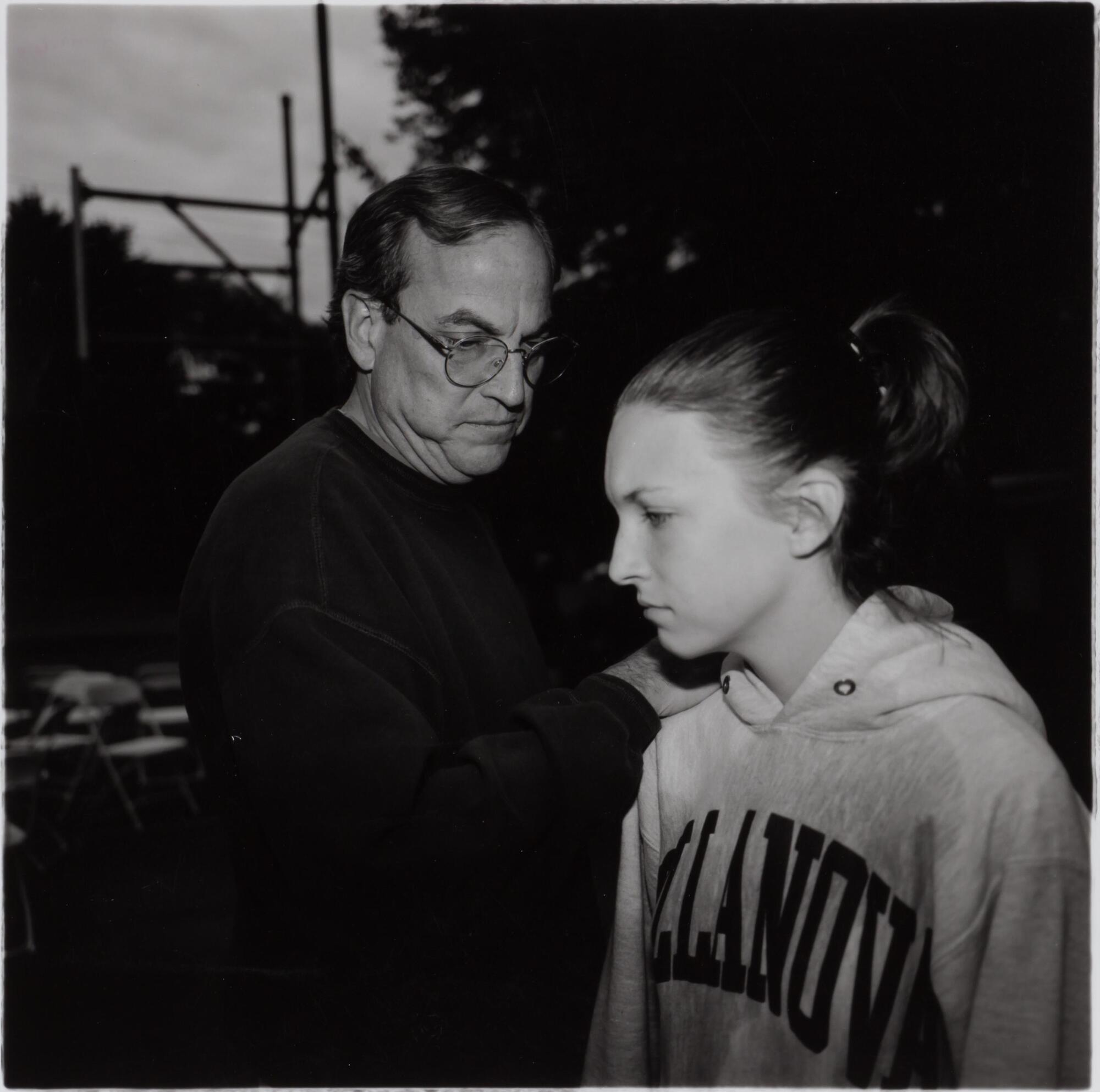 A photograph of a man wearing a dark shirt and glasses,and a young woman wearing a sports sweat shirt. The man turns toward the girl, placing his hand on her shoulder. Both have solemn expressions.