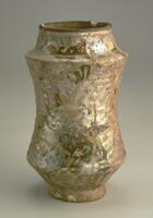 A 14th century Mongol period pharmacy jar from the Sultanabad region. The albarello is a contracted cylinder with a converging neck and has a conventionalized inscription with a stag and gazelle design. Floral designs fill the space while the lower body is fluted. All the decorative patterns were done in low relief. Paste is a light tan "Islamic II", while the glaze is glossy with fine crackle. The object was fired upright. Colors include heavy iridescent patina, gray-green, light grayish-white ground and light purplish gray. The object has been restored. 