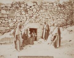In this photograph, eleven figures—seven adults and three children—stand and sit near a portal set into a stone wall. 