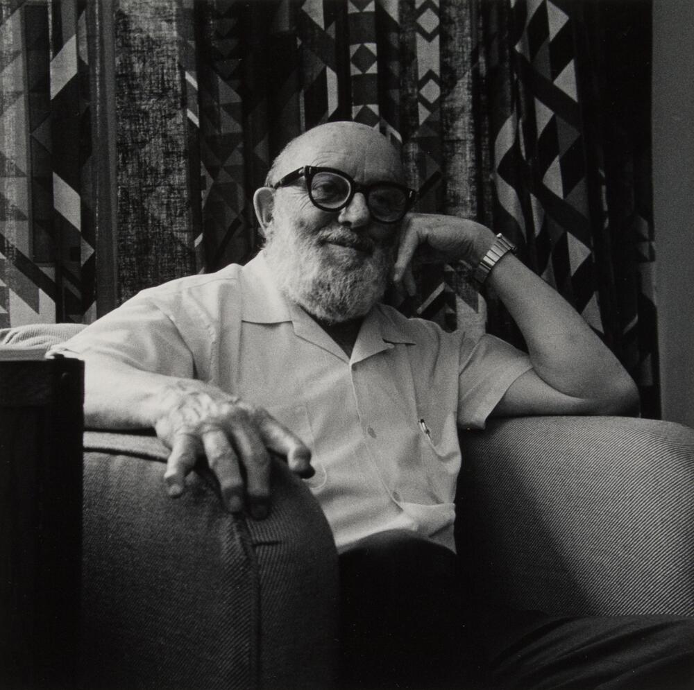 Ansel Adams seated in a chair, graphic curtains behind. His hand is to his head and he is wearing glasses.