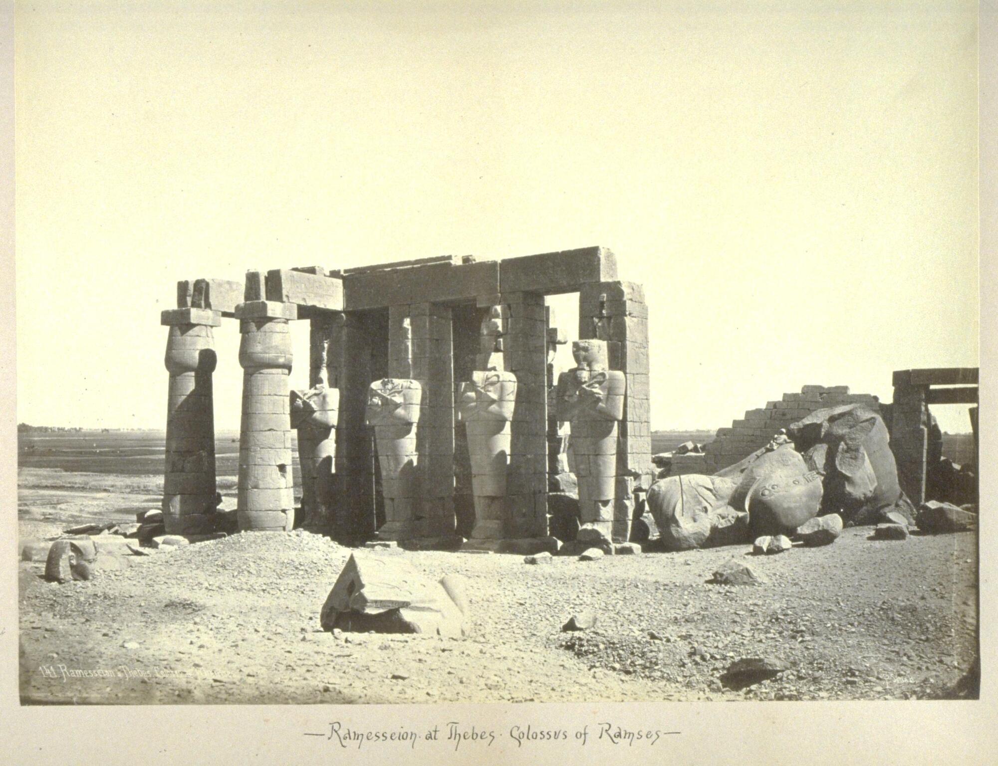 A grouping of columns and figural sculptures stands amid other ruins and stone debris in a mostly flat, rocky landscape. 