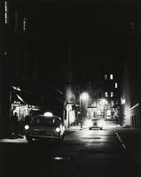 Black and white image of a night scene of a city street with a taxi pulled to the side of the curb, another coming towards it and with lights on.