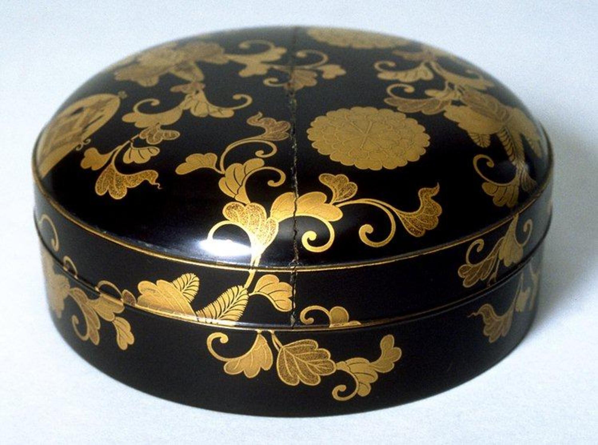 A short, round container lacquered with gold floral patterns wrapping around the entire container. A small crysanthemum crest and geometric crests, also in gold, appear on the rounded top of the container. Part of a bridal trousseau.