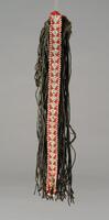 An apron in the form of a dark colored string fringe attached to a narrow strip of red and white beadwork with a triangular design. Small, white buttons hang from the bottom of the beadwork. 